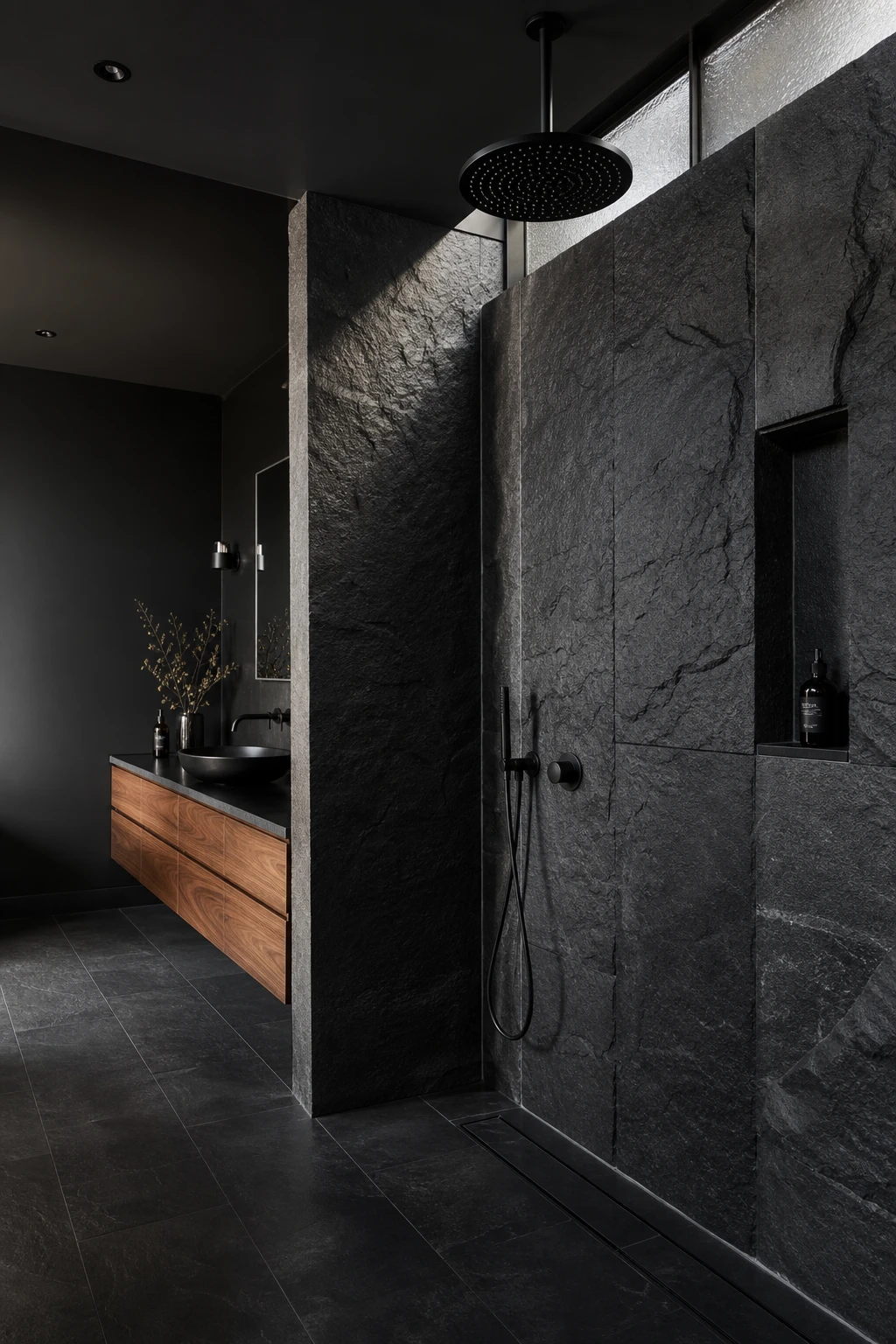 Luxury black bathroom with a black stone shower as the hero feature, raw textured walls, matte black fixtures, and warm directional lighting on dark natural stone