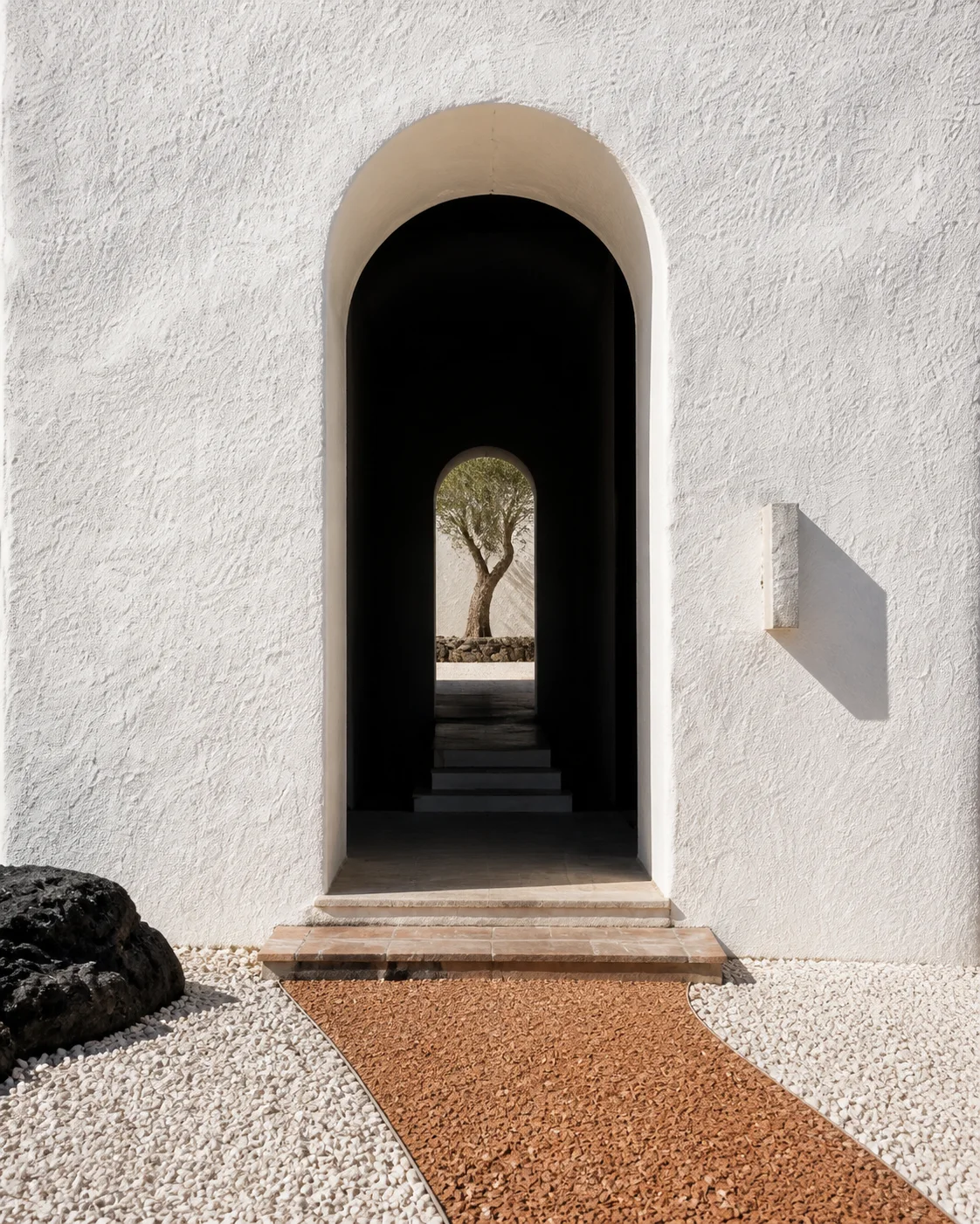 Deep black arched threshold set in chalk white lime plaster with terracotta steps pale aggregate volcanic rock and strong southern sunlight in the Milos house part of an editorial.