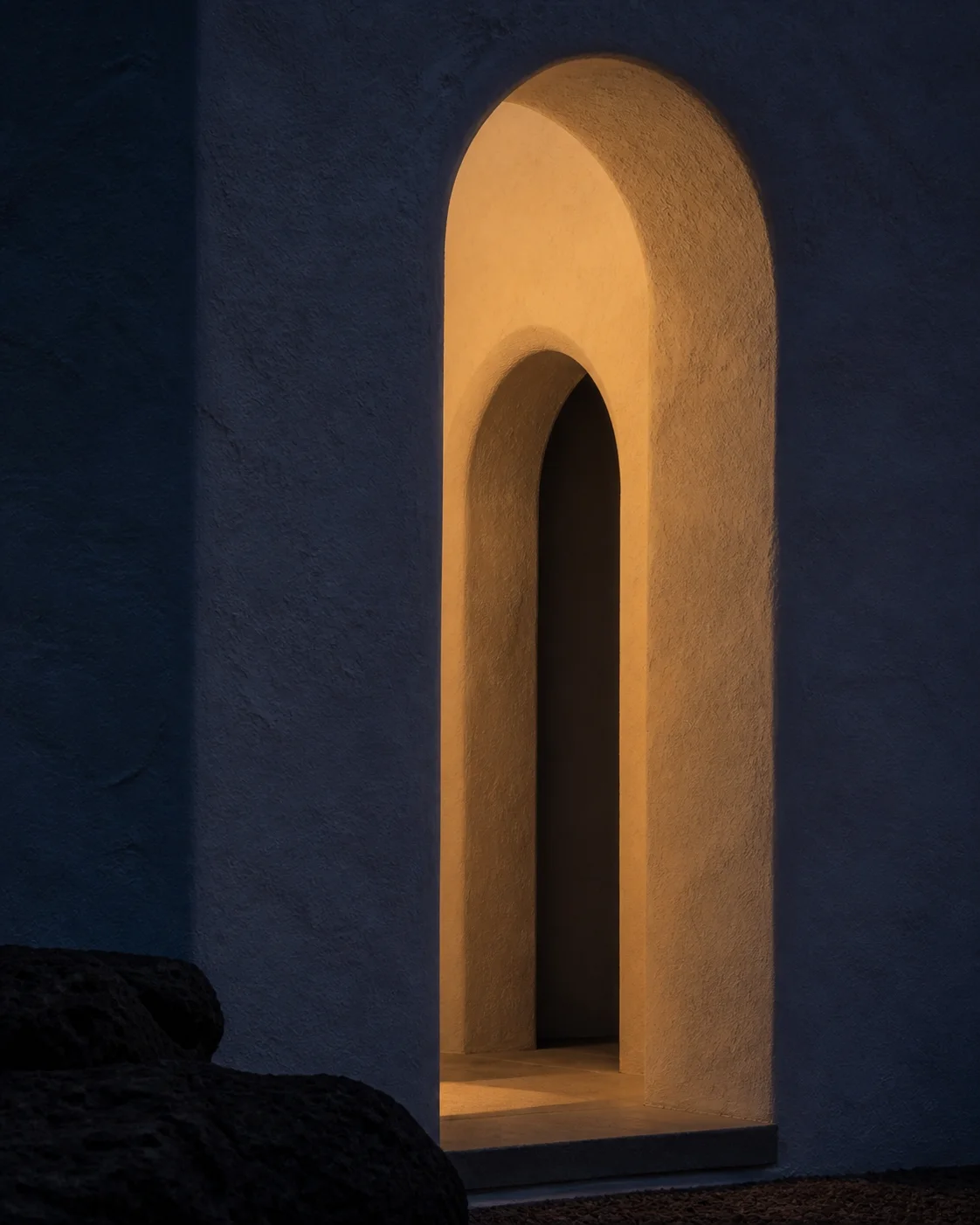 Intimate dusk detail of a dark plaster arch with restrained warm interior glow volcanic shadow and deep cobalt evening light in Milos part of an editorial architecture walkthrough.