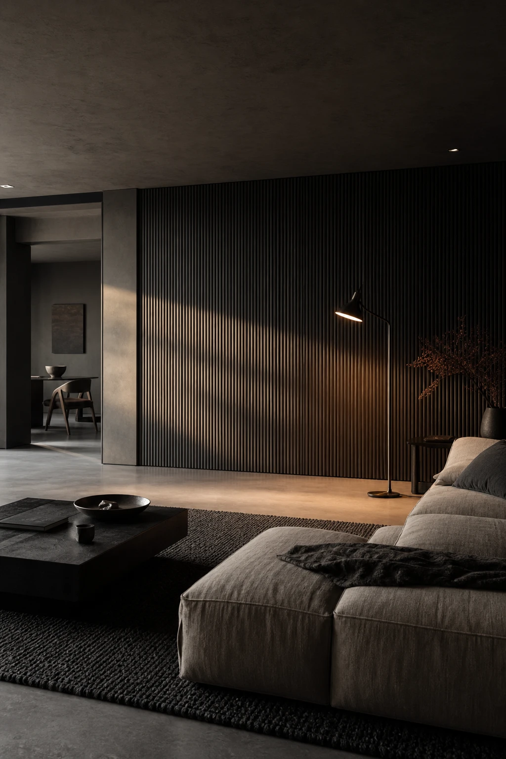 Dark minimalist living room with tonal layering in charcoal, slate and warm black, featuring a low linen sofa, ribbed plaster walls and soft evening light