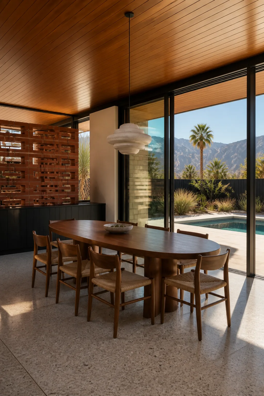 A walnut dining table with woven chairs, a travertine pendant and pool view in the Palm Springs house.