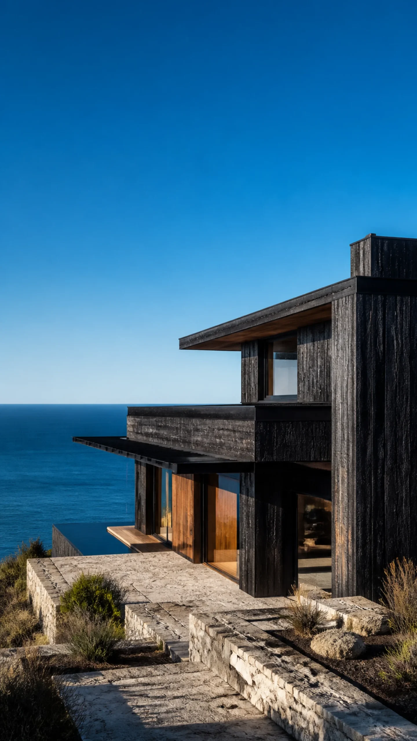 Whole black shou sugi ban coastal house above pale limestone terraces and a deep blue Aegean sea An original fictional editorial architecture study of material landscape light and.